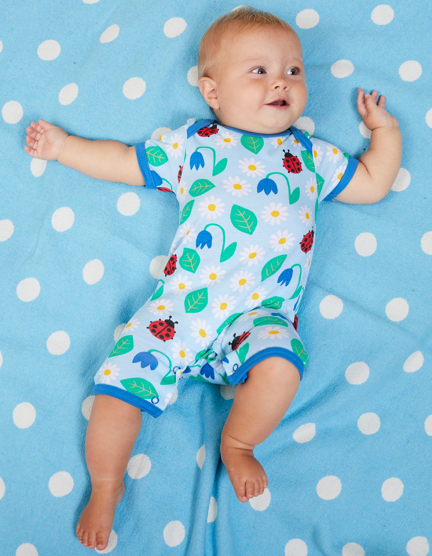 Žaismingas vaikiškas romperis trumpomis rankovėmis ir šortukais, šviesiai mėlynas, margintas boružėlėmis ir smulkiomis gėlėmis.